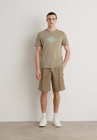 Tan t-skjorte med grønn "HUGO" logo, parret med tan shorts. Hvite sneakers og sokker. Glatt tekstur, uformelt design, avslappet passform.