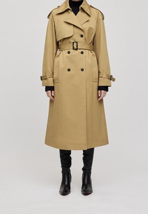 Gabardina beige de doble botonadura con cinturón, hombreras y correas en los puños, llevada sobre un jersey de cuello alto negro y botas negras hasta la rodilla.