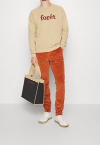 Persona vistiendo un sudadero beige con el texto "forét", pantalones de pana color óxido, zapatillas blancas, sosteniendo un bolso tote de Marni en negro y beige.