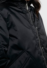Giacca imbottita nera con colletto alto, zip argentata e cuciture a vista. Include cordoncini e una tasca frontale con patta triangolare.