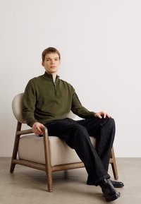 Olivgrüner Pullover mit Viertelreißverschluss, dunkle Jeans, schwarze Knöchelstiefel, sitzt auf einem beigen Stuhl, neutraler Hintergrund.