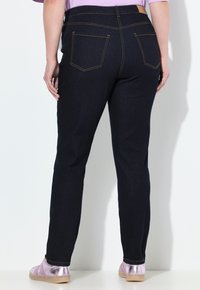 Tmavě modré džíny z denimu s elastickým pasem střední výšky, které mají dvě zadní kapsy a kontrastní žluté stehy. Nosené s světle růžovými botami.