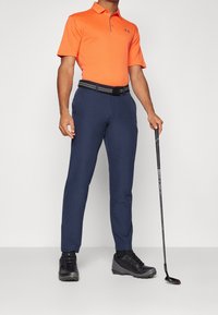 Oranžová polo košile s krátkým rukávem, námořnické kalhoty, černý opasek a černé golfové boty. Drží golfovou hůl, stojí na neutrálním pozadí.