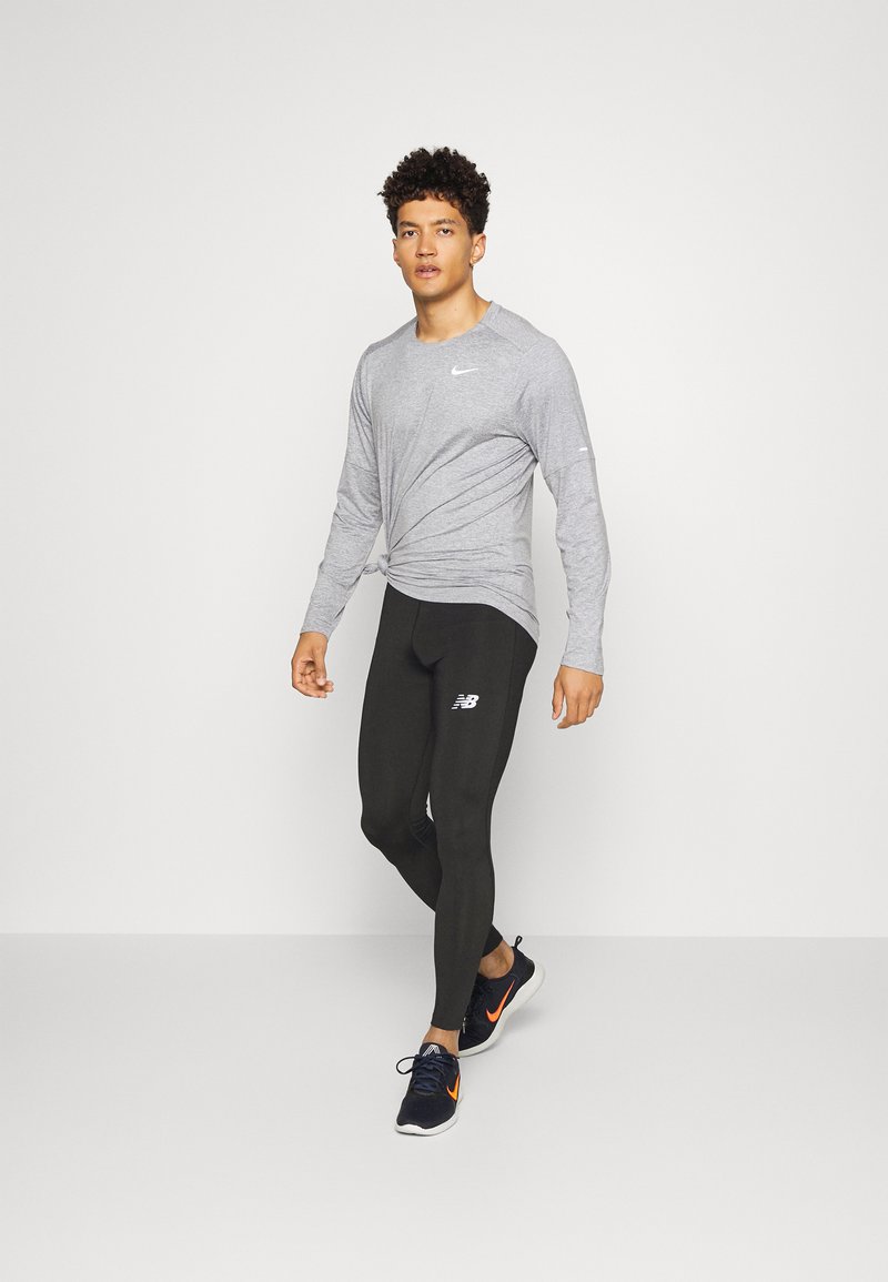 Hombre con camiseta de manga larga gris, leggings negros y zapatillas negras con un swoosh naranja caminando sobre un fondo blanco liso.