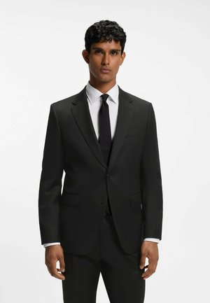 Uomo che indossa un abito nero su misura con camicia bianca e cravatta nera, in piedi davanti a uno sfondo bianco uniforme.