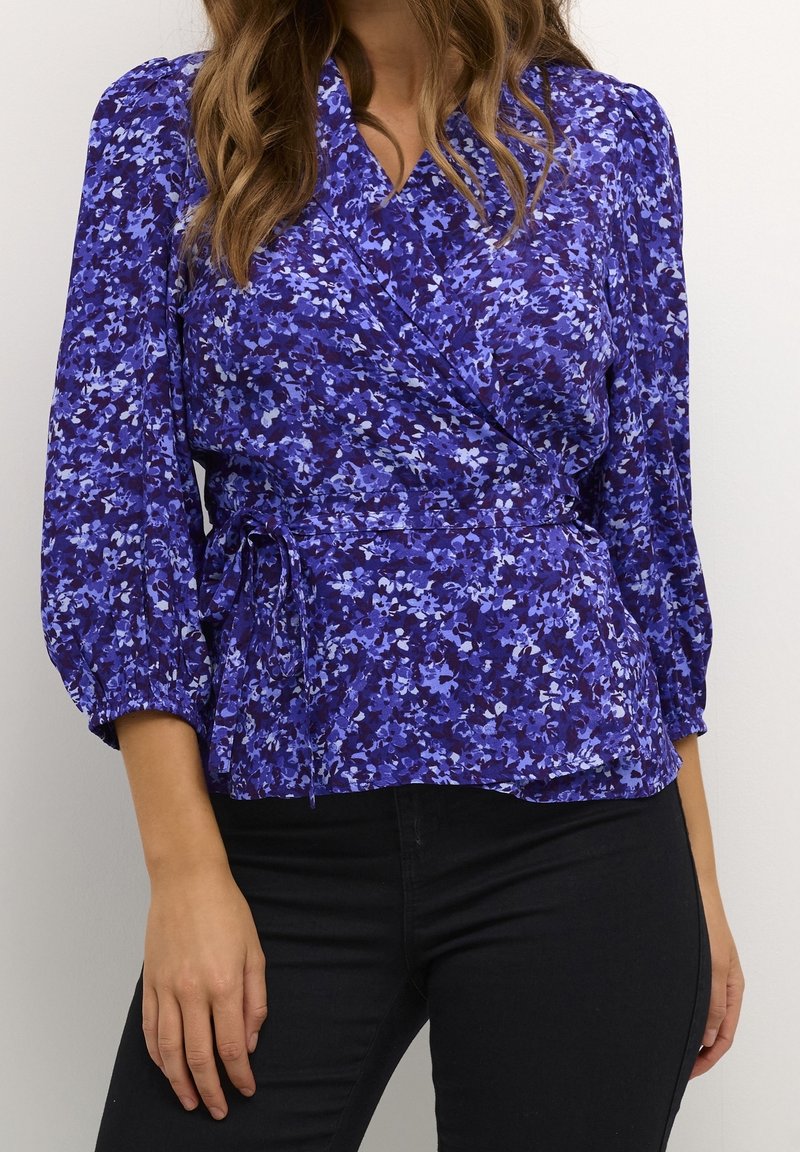 Blouse portefeuille en tissu violet avec un motif floral dans des tons de bleu. Manches longues et ceinture à nouer à la taille pour un ajustement personnalisé.