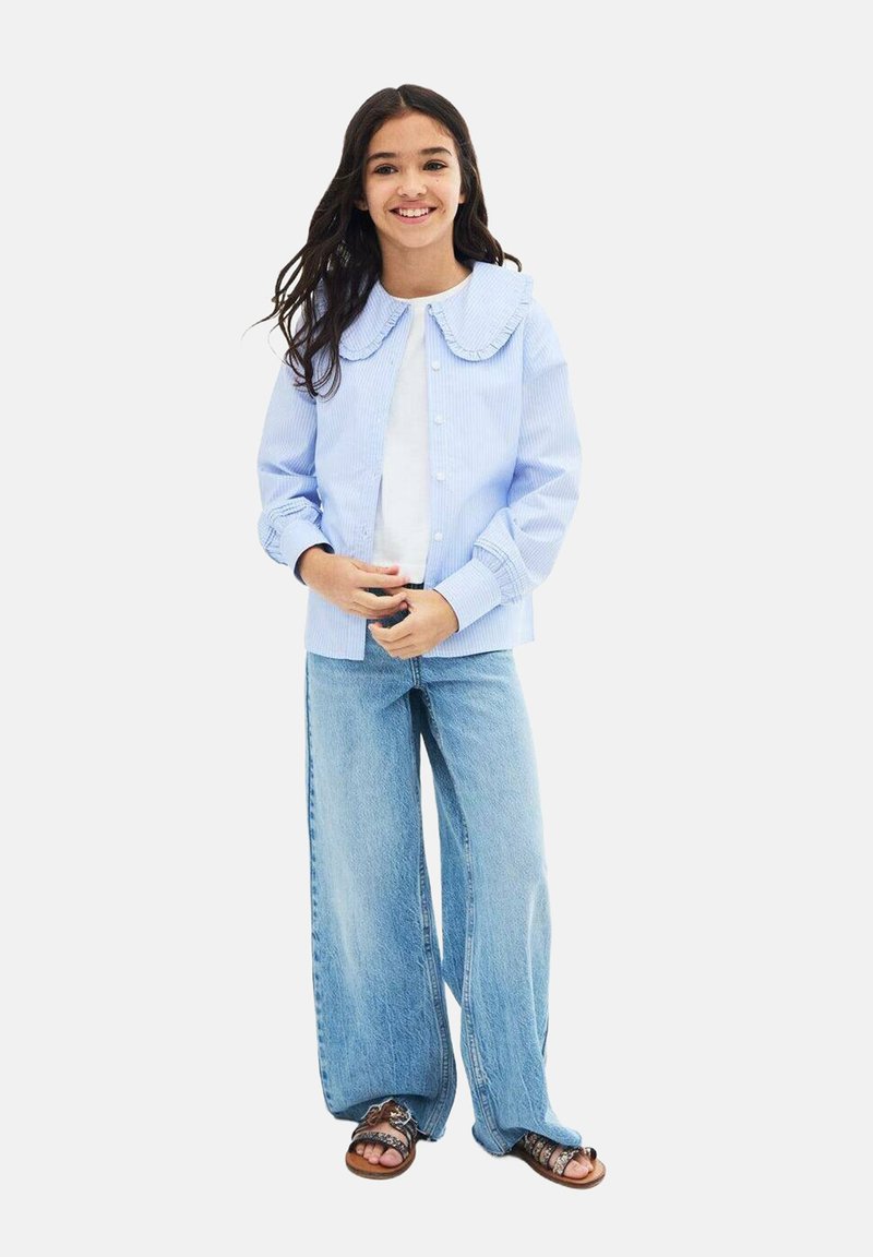 Vaaleansininen raidallinen takki, jossa on suuri rypytetty kaulus, yhdistetty leveälahkeisiin farkkuihin ja ruskeisiin sandaaleihin. Valkoinen t-paita alla.