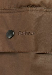 Barna szövetkabát sima textúrával, fekete gombbal és hímzett "Barbour" logóval, a minimalista dizájnt kiemelve.