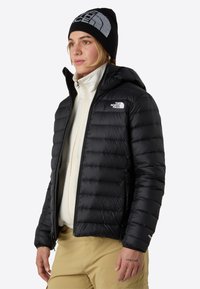 Svart puffjacka med huva, quiltad design och dragkedjestängning. Bärs över en ljusfärgad fleece, har logotyp på bröstet och ärmarna.