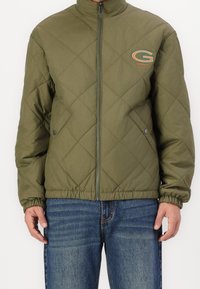 Olivengrøn quiltet jakke med høj krave, frontlynlås og elastisk søm. Har et farverigt GANT-logo på venstre bryst.