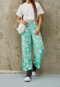 Bílá zkrácená košile na knoflíky, tyrkysové květinové široké kalhoty a černé tenisky s bílými akcenty. Doplňky zahrnují malou bílou tašku.