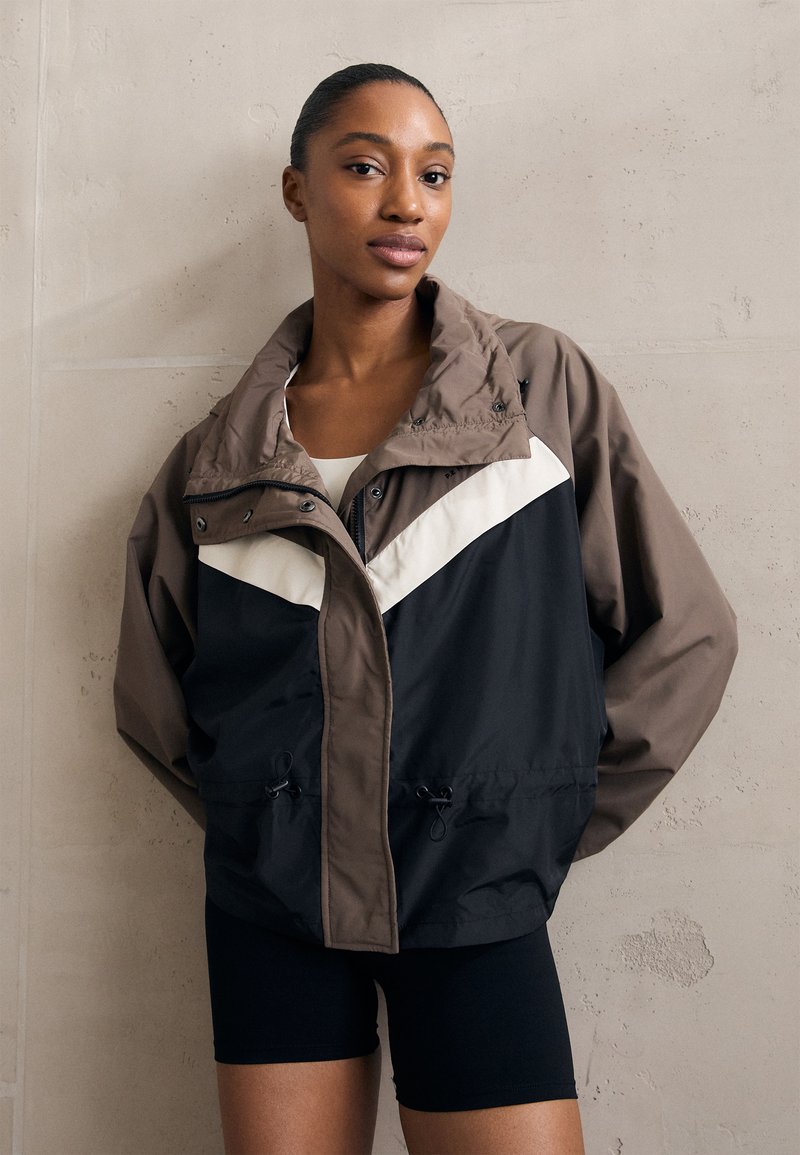 Braune und schwarze Windjacke mit einem weißen diagonalen Streifen, verstellbaren Kordeln am Saum und einem Stehkragen.