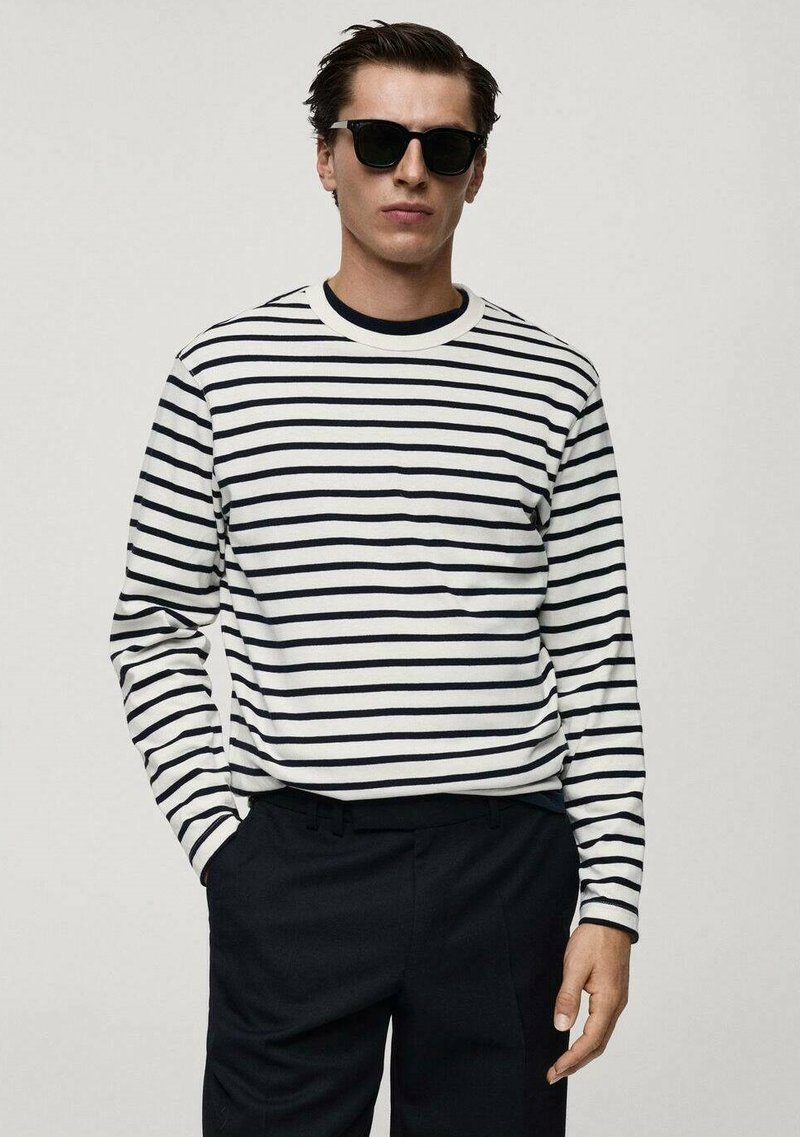 Langärmliges gestreiftes Hemd in Weiß und Marineblau mit Rundhalsausschnitt. Das Model trägt schwarze Sonnenbrille mit gerade geschnittenen Hosen und die Hände in den Taschen.