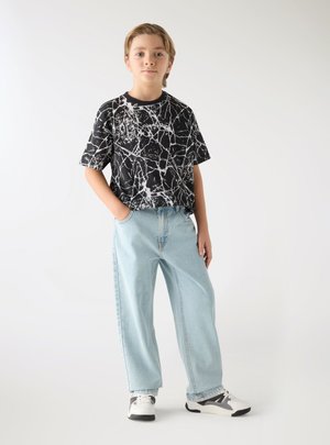 Giovane persona in piedi con le mani nelle tasche, che indossa una camicia a fantasia bianco e nero, jeans azzurri e sneakers bianche.