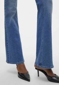 Modri kavbojki s širokimi hlačnicami in vidnimi šivi, v kombinaciji s črnimi lakastimi čevlji z ostrim nosom. Robovi imajo rahlo strganje.