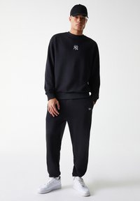 Sudadera negra con logo visible, pantalones joggers negros y zapatillas blancas. Tejido suave, corte relajado y puños elásticos. Gorro de béisbol negro.