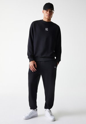 Sudadera negra con logo visible, pantalones joggers negros y zapatillas blancas. Tejido suave, corte relajado y puños elásticos. Gorro de béisbol negro.