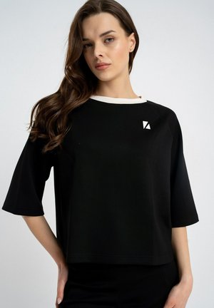 Kobieta z długimi brązowymi włosami, ubrana w czarną bluzkę z krótkim rękawem, białym kołnierzykiem i małym białym geometrycznym logo na piersi, na jednolitym tle.