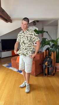 Man glimlacht en staat binnen, draagt een patroon kortemouwenoverhemd, lichte korte broek en witte sneakers in een moderne woonkamer.