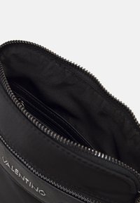 Černá nylonová taška se zipovým uzávěrem, s hladkým povrchem a malou vnitřní kapsou. Obsahuje logo na předním panelu.