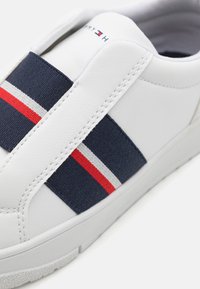 Sneaker alb din piele cu brida din textil bleumarin care prezintă dungi roșii și albe. Textură netedă și detalii de coasere subtile.