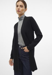 Cardigan nero, design con apertura frontale, texture morbida, maniche lunghe, abbinato a un dolcevita grigio e denim scuro. Stile minimalista.