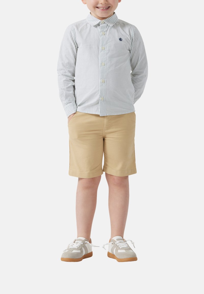 Bambino con una camicia a righe azzurro chiaro e bianca, pantaloni corti beige e sneakers grigie con lacci bianchi e suole in gomma.
