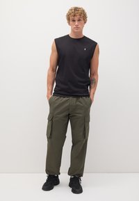 Maglietta nera senza maniche con un piccolo logo, abbinata a pantaloni cargo verde oliva con più tasche e sneakers nere.