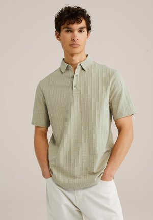 Short-sleeve polo shirt in light green with a textured pattern, featuring a collar, button placket, and relaxed fit. Worn with light-colored pants.