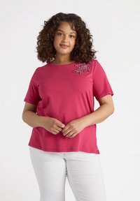 Camiseta de manga corta en fucsia, hecha de tela suave, con un diseño floral de lentejuelas en el hombro izquierdo y un cuello redondo.