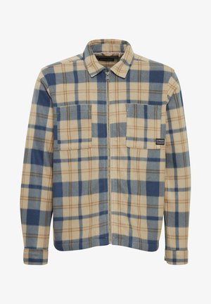 Camicia in flanella a scacchi beige e blu, con chiusura a zip, due tasche sul petto e una consistenza morbida. Linee orizzontali e verticali creano un motivo.