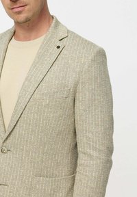 Egy férfi bézs textúrájú zakót visel világos bézs ing felett, a felső teste és az arca egy része látható egy fehéren.