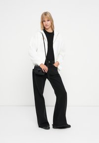 Hvit hettejakke over en svart topp, kombinert med svarte vide jeans og svarte sko. En liten svart crossbody-veske fullfører antrekket.