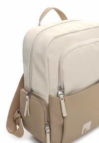 Krémový a béžový batoh z hladké syntetické látky. Má několik kapes na zip a horní držadlo pro snadné nošení.