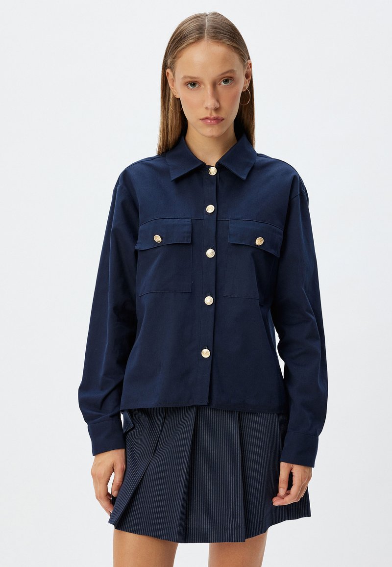 Camicia blu navy con bottoni in un tessuto morbido, dotata di due tasche frontali, bottoni dorati e una vestibilità rilassata, abbinata a una gonna plissettata.