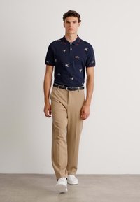 Navy poloskjorte med fuglemønster, røde krageaksenter; beige bukser; modellen har på seg hvite joggesko og står på en nøytral bakgrunn.