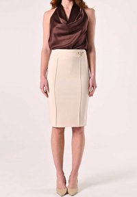 Femme portant un haut satiné sans manches marron rentré dans une jupe crayon beige taille haute avec des détails en boutons dorés, associée à des escarpins beiges.
