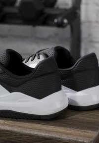 Chaussures de sport noires et blanches en tissu texturé posées sur une surface en bois, équipement de gym flou en arrière-plan.