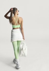 Mintgroene sportbeha met kruisende bandjes, witte omslagrok, bijpassende groene legging en een witte tas. Model draagt witte sneakers.