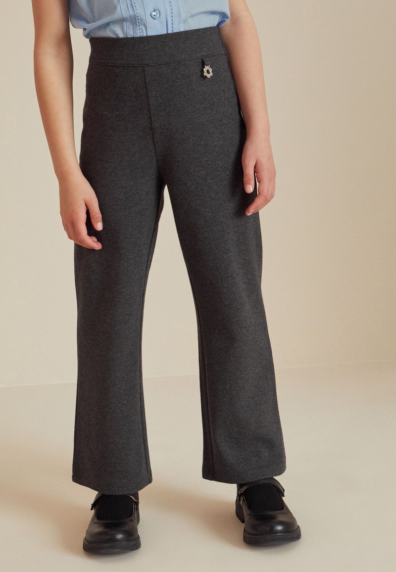 Enfant portant un pantalon gris foncé avec un détail bouton fleur, chaussures noires et chemise bleu clair boutonnée, debout sur un sol beige.