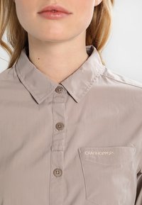 Camicia button-up color beige chiaro con colletto, dotata di bottoni marroni, taschino sul petto e logo ricamato "Craghoppers". Texture liscia.