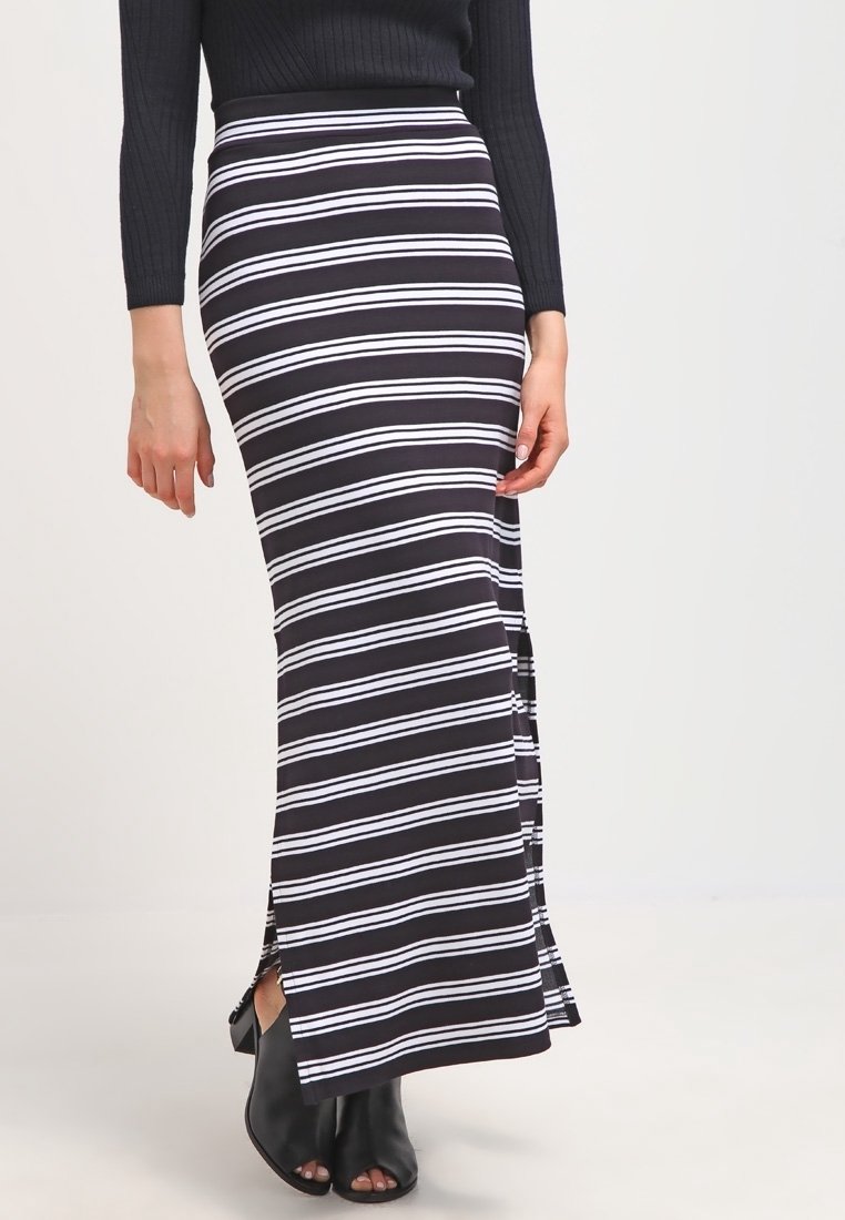 Jupe maxi à rayures noires et blanches avec une taille ajustée et des fentes latérales, fabriquée dans un matériau stretch doux. Associée à des chaussures ouvertes noires.