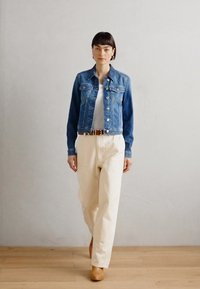 Giacca di jeans con bottoni argentati, top bianco e pantaloni a gamba larga color crema. Cintura marrone e stivali beige. Sfondo neutro e pavimento in legno.