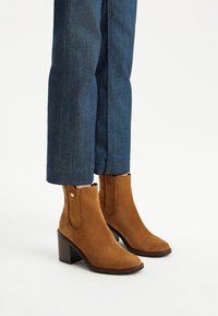 Brązowe zamszowe botki za kostkę z rzeźbioną drewnian�ą obcasem, z elastycznymi bocznymi panelami i gładką strukturą. Noszone z ciemnoniebieskimi, skróconymi jeansami.