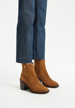 Brązowe zamszowe botki za kostkę z rzeźbioną drewnianą obcasem, z elastycznymi bocznymi panelami i gładką strukturą. Noszone z ciemnoniebieskimi, skróconymi jeansami.