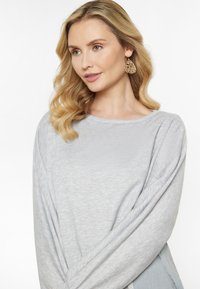 Lys grå langærmet top med rund halsudskæring og blød tekstur. Modellen bærer detaljerede guldøreringe, som viser et afslappet look.