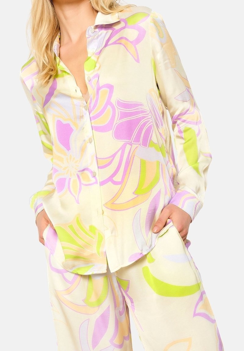 Žena v volné saténové košili a kalhotách s velkými pastelovými květinovými vzory v purpurové, žluté a zelené barvě, s rukama v kapsách.
