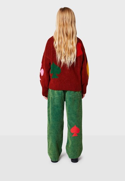 Červený sveter s viacfarebnými geometrickými vzormi, kombinovaný so zelenými kordrovými nohavicami s červeným motívom piky. Dlhé vlasy sú viditeľné.