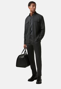 Tmavě šedá textilní košile se dvěma kapsami na hrudi, spárovaná s odpovídajícími kalhotami a černými koženými botami. Černá kožená taška přes rameno držena v ruce.