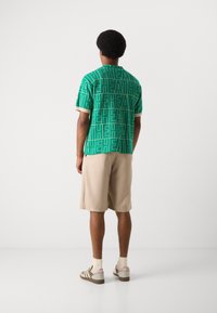 Grøn kortærmet skjorte med stort "HEATH" print, creme kant, parret med beige shorts og multifarvet fodtøj.